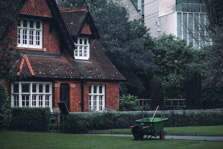 A traditional red-brick house with a pitched, tiled roof and white-framed sash windows, set within a well-maintained garden. The garden features lush green lawns, neatly trimmed hedges, and a border o