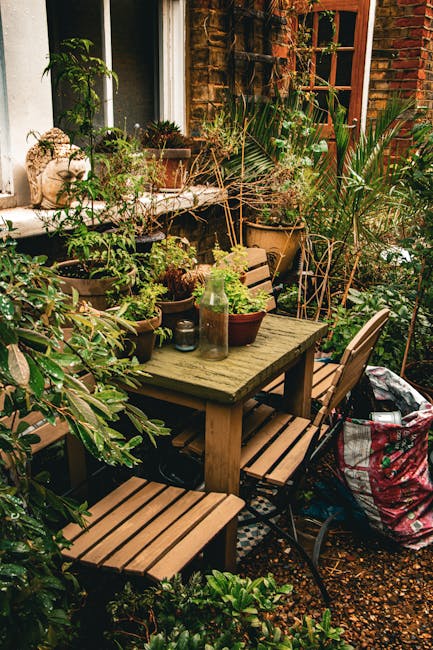 A cozy outdoor garden area adjacent to a brick house, featuring a weathered wooden table surrounded by matching wooden chairs, all set on a gravel surface. The garden is lush with various potted plant